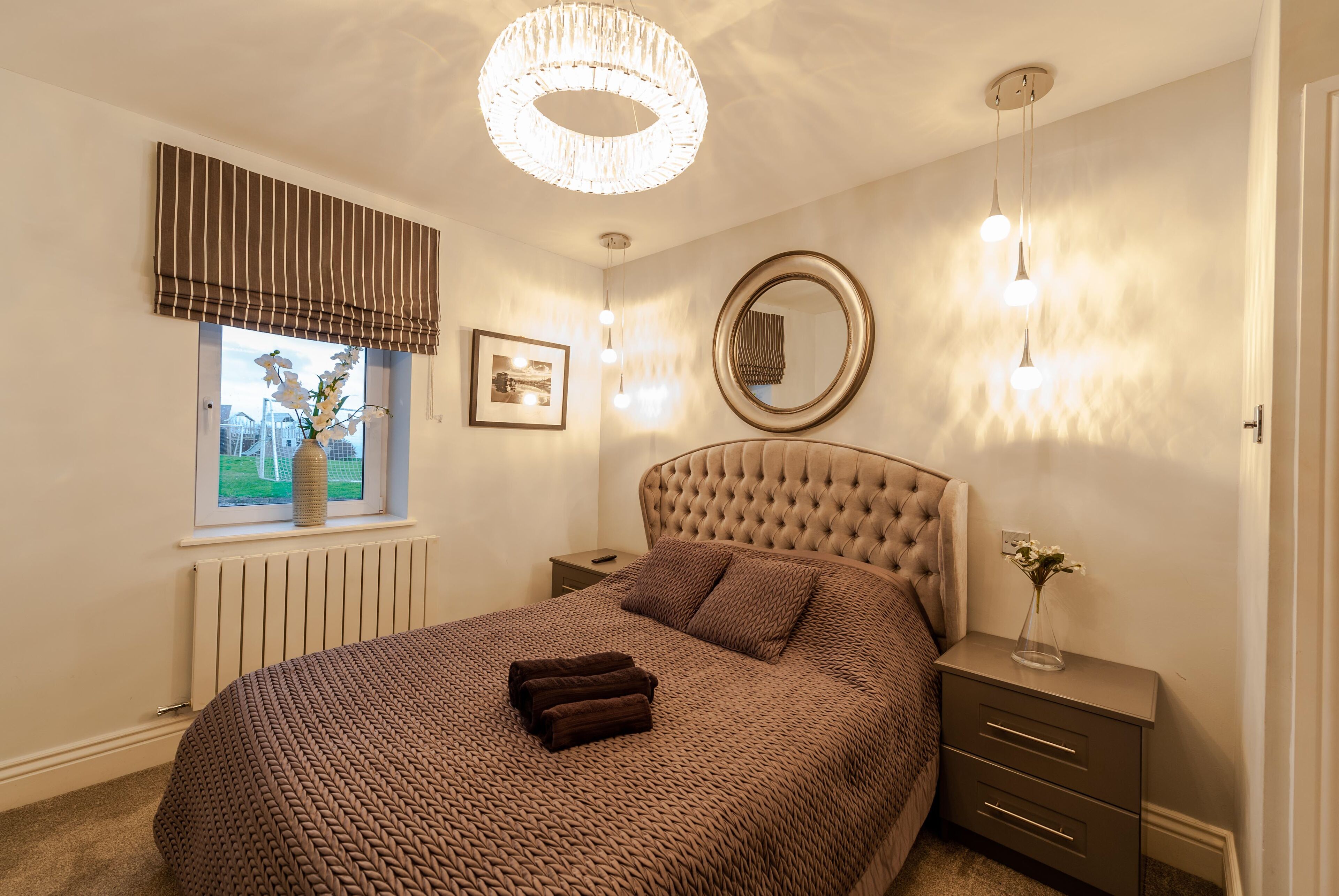 Un dormitorio con una cama grande, una ventana con cortinas, una mesita de noche con lámpara y un espejo en la pared.