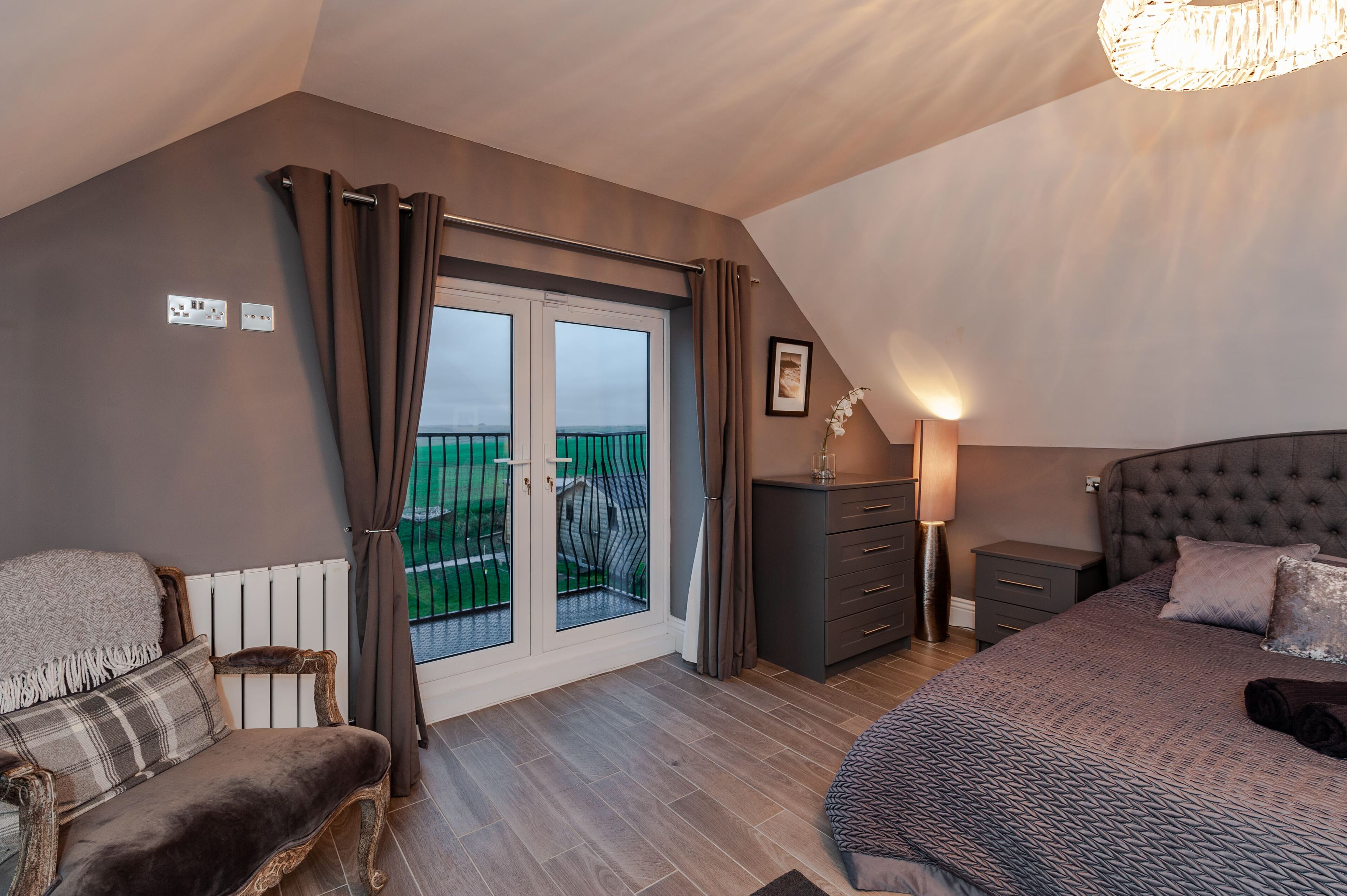A bedroom with a large bed, a dresser, a nightstand, a chair, and a view of a balcony.