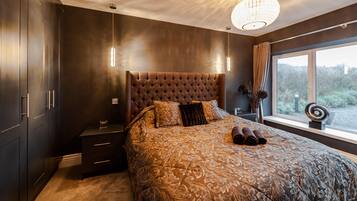A bedroom with a tufted headboard, a large bed with patterned bedding, bedside tables, a nightstand, a window with a view, and a hanging light fixture.