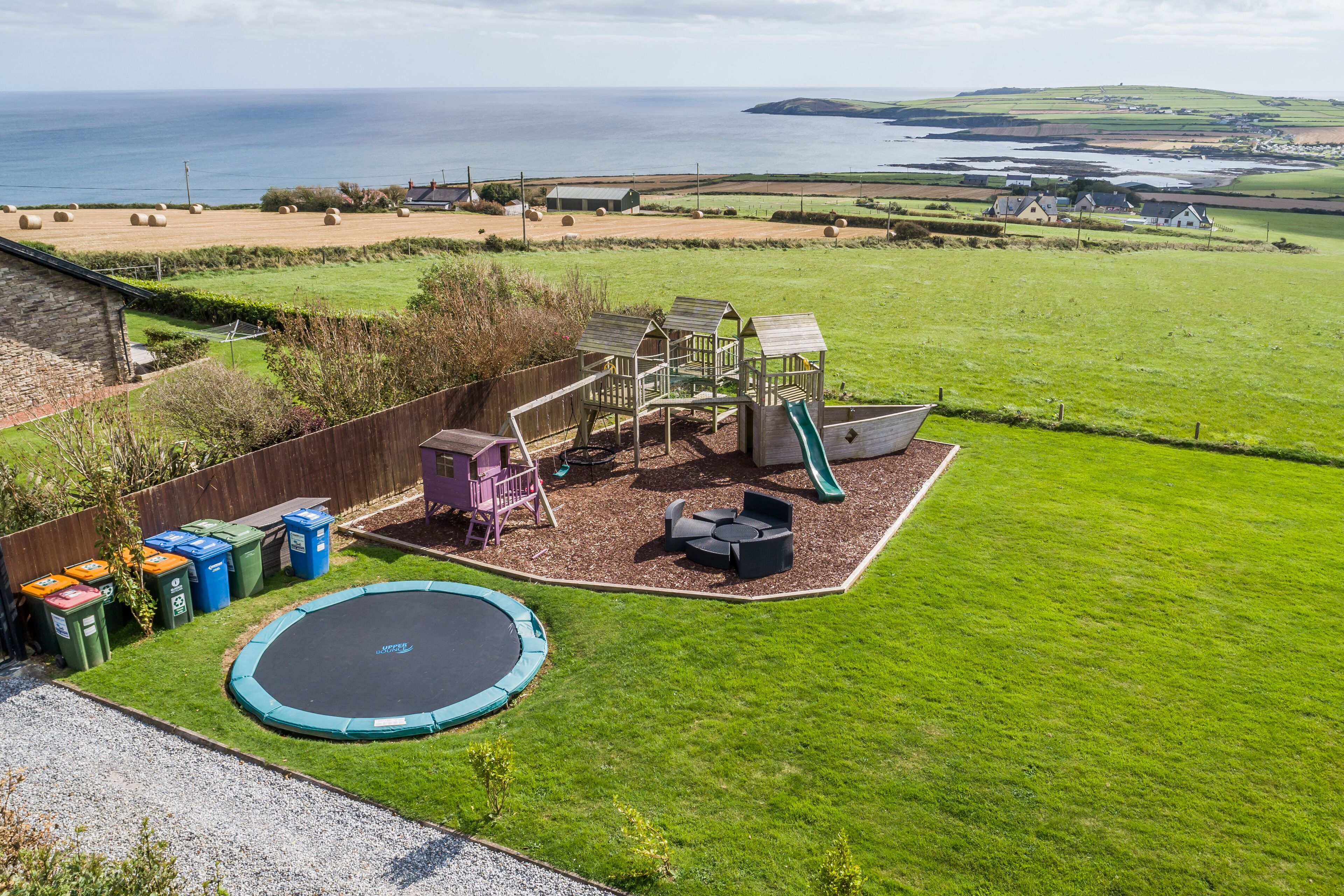 A playground with a slide, swings, and a trampoline in a grassy area with a view of the sea.