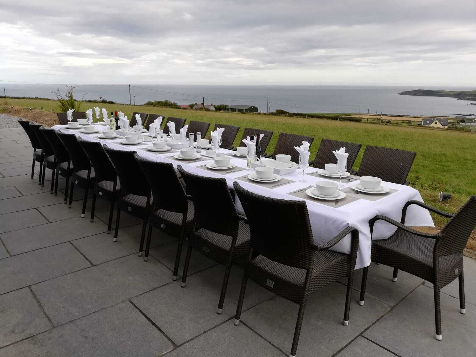 A long dining table set outdoors with chairs, overlooking a coastal landscape.