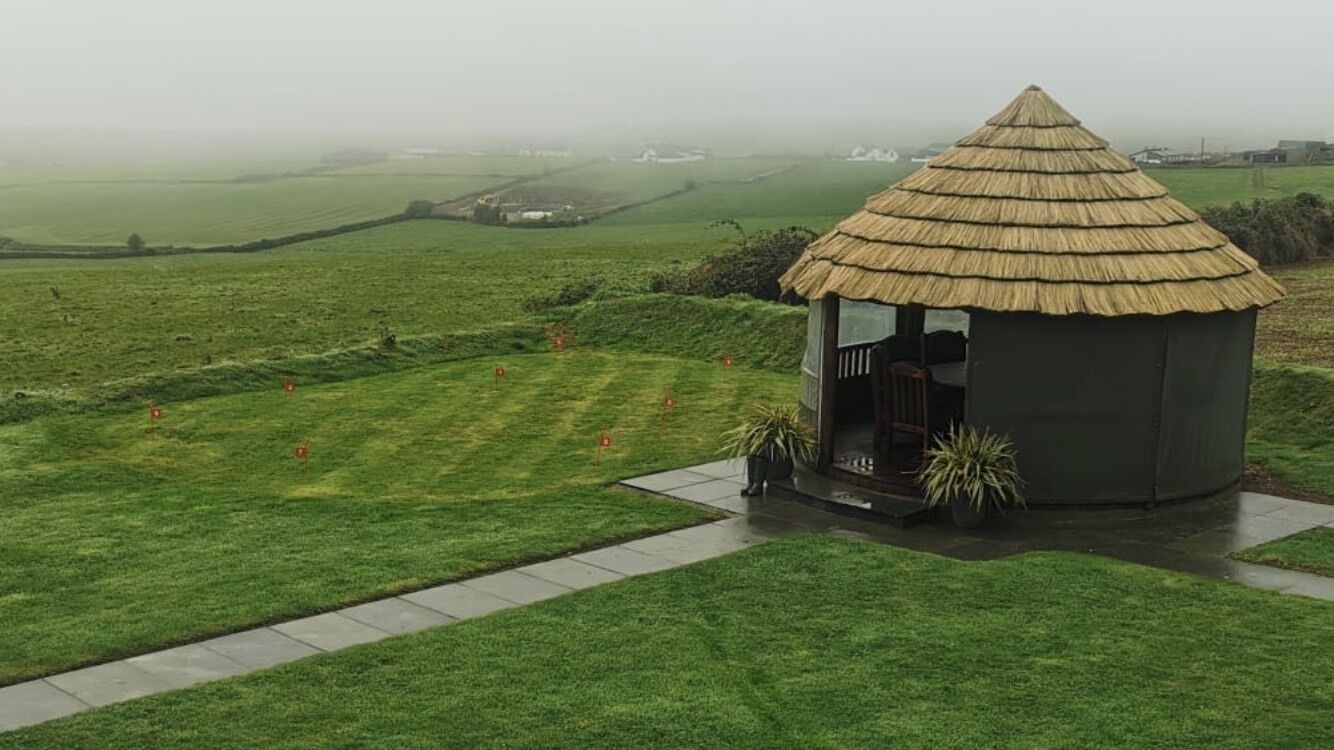 Uma construção com telhado de palha e uma pequena varanda, cercada por um gramado bem cuidado e uma paisagem rural ao fundo.