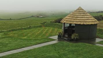 Uma construção com telhado de palha e uma pequena varanda, cercada por um gramado bem cuidado e uma paisagem rural ao fundo.