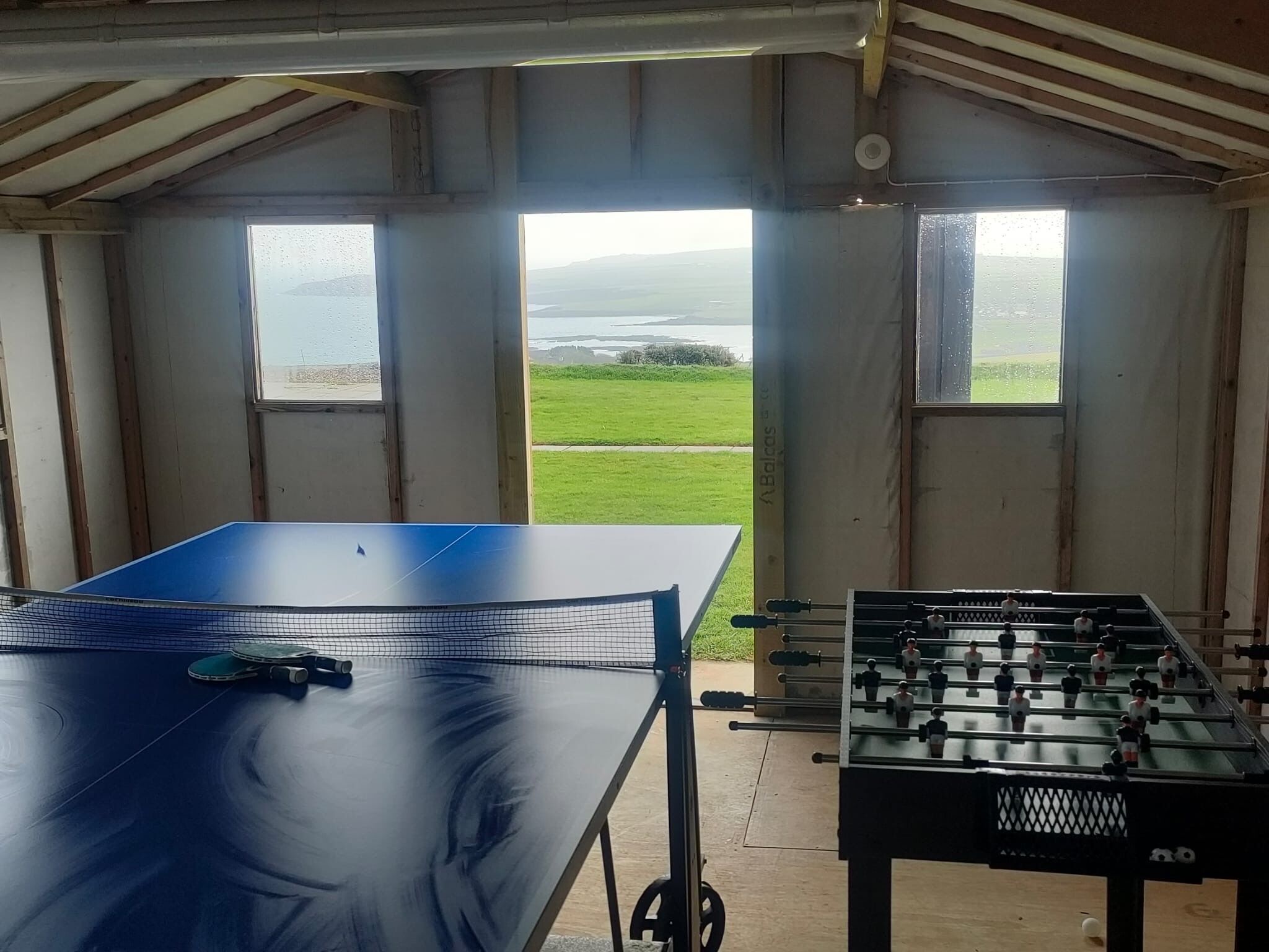 Ein Zimmer mit Tischtennisplatte und Kicker, große Fenster mit Blick auf ein grünes Feld und Wasser.