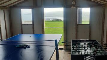 Ein Zimmer mit Tischtennisplatte und Kicker, große Fenster mit Blick auf ein grünes Feld und Wasser.