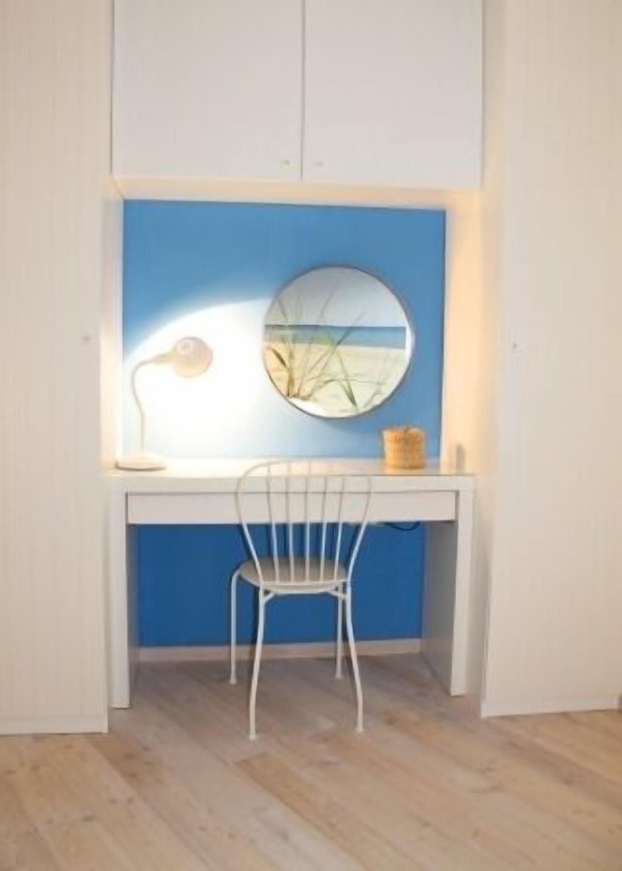Una habitación con un escritorio, una silla, una lámpara y un espejo redondo que muestra una escena de playa.