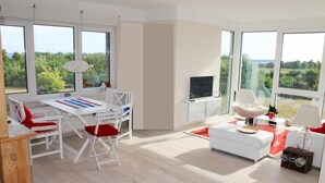 A modern living room with a dining area, large windows, and a view of greenery. - Light-flooded apartment with private sauna! Beach walking distance, with dog! (Hohenkirchen)