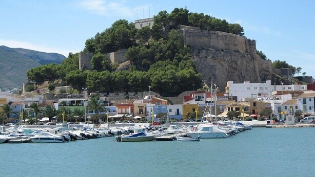 A coastal town with a historic fort on a hill, a marina with boats, and colorful buildings.