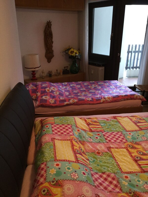 A room with two beds covered in colorful quilts, a wooden nightstand with a lamp and a vase of flowers, and a view of a wooden fence outside.