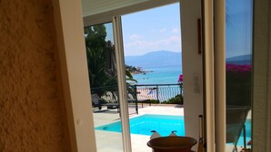Vista a través de un ventanal hacia una zona de piscina con un jarrón grande, una silla y vistas al mar y a las montañas.