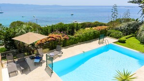 Una piscina con vistas al mar, rodeada de vegetación y un patio con sillas y una sombrilla.