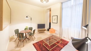 A modern living room with a round dining table, comfortable seating, a flat-screen TV, and a large window with sheer curtains.