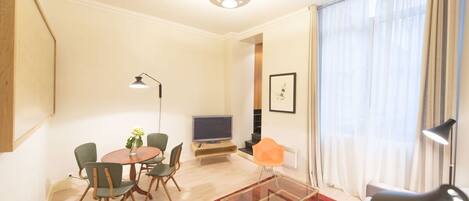 A modern living room with a round dining table, comfortable seating, a flat-screen TV, and a large window with sheer curtains.