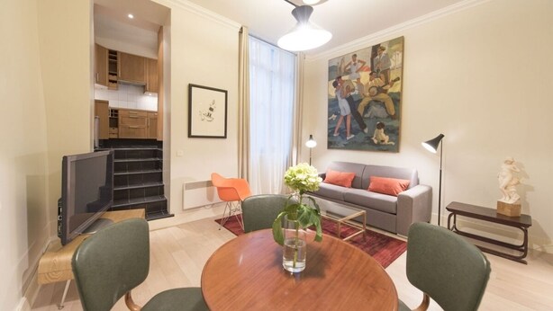 A modern living room with a dining table, green chairs, a grey sofa, and a painting on the wall.