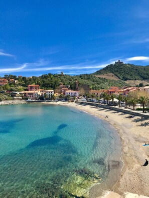 A coastal town with a clear beach, buildings with red roofs, and a hill with a windmill.