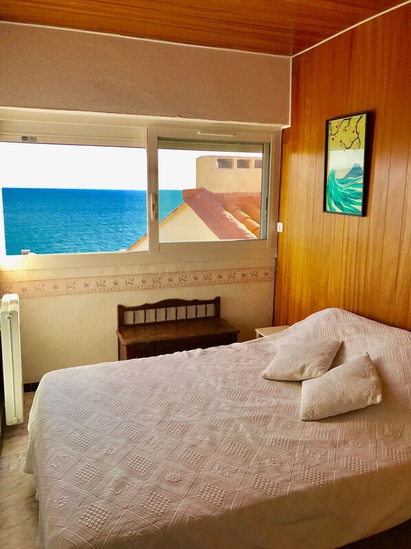 A bedroom with a bed, a wooden headboard, a nightstand, a window with a view of the sea, and a painting on the wall.