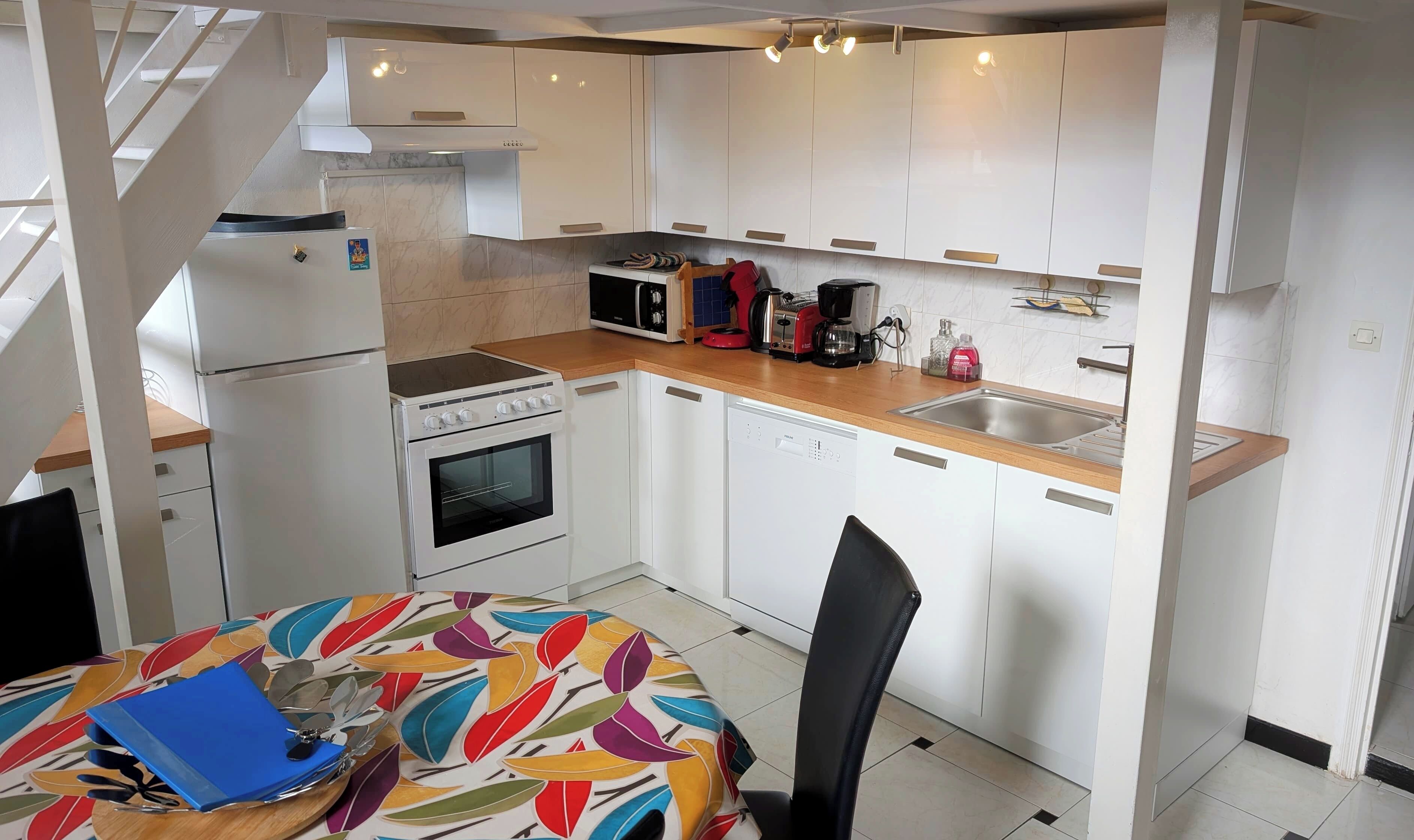 Une cuisine compacte avec des meubles blancs, un four à micro-ondes, une cuisinière et un lave-vaisselle. Elle est dotée d’un plan de travail en bois et d’un petit évier.