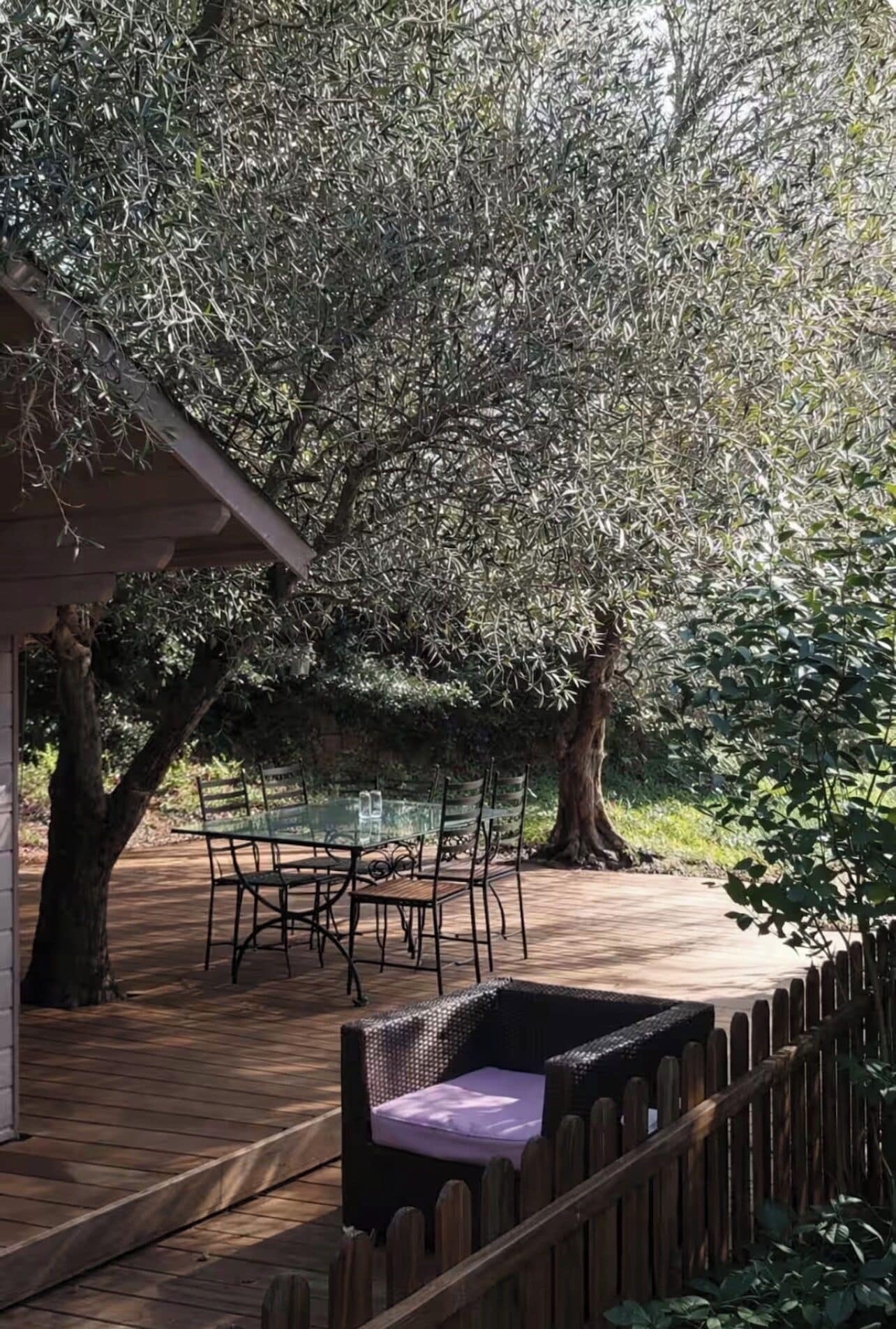 An outdoor patio with a glass table and chairs, surrounded by trees and a wooden fence.