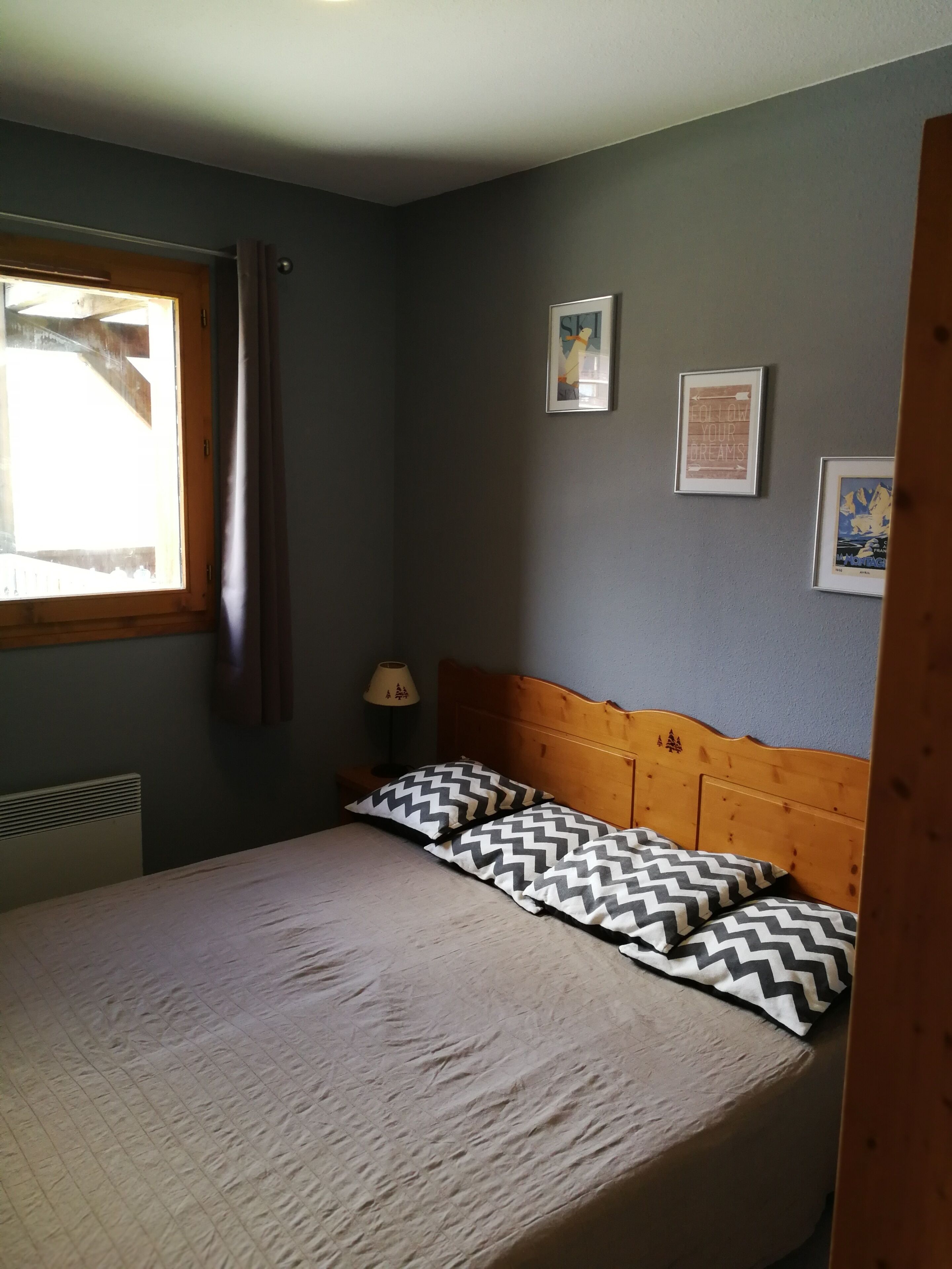 Een slaapkamer met een bed, een raam met gordijnen, een nachtkastje met een lamp en twee ingelijste foto's aan de muur.