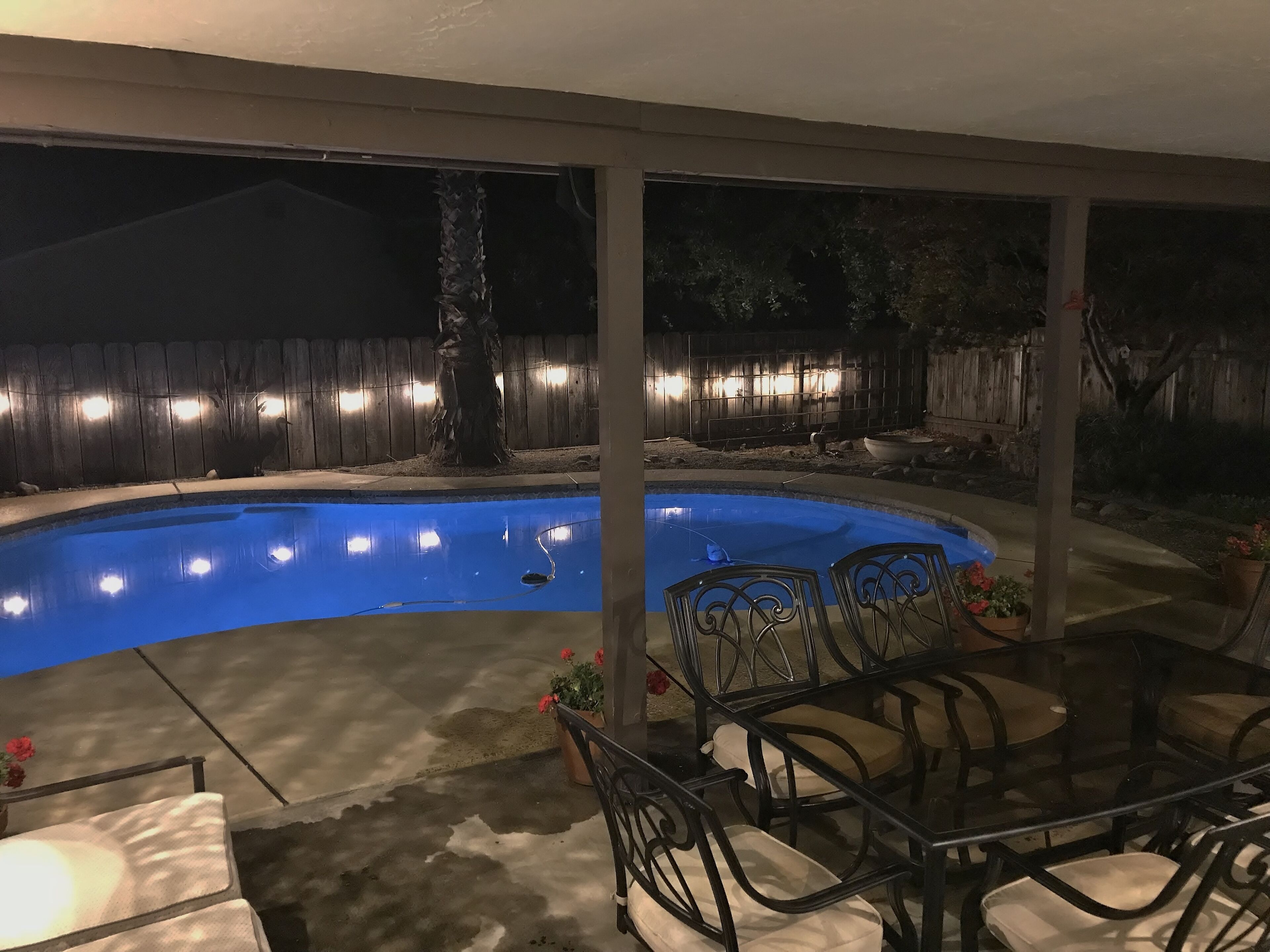 A swimming pool with a curved edge, surrounded by a covered patio area with metal chairs and tables.