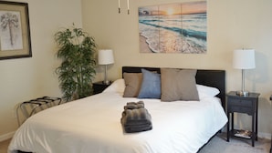 A neatly made bed with a folded towel, a nightstand with a lamp, a potted plant, and a painting of a beach scene. - The Pineapple House -Best Reviews- Refreshing Swimming Pool! (Redding)