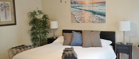 A neatly made bed with a folded towel, a nightstand with a lamp, a potted plant, and a painting of a beach scene.