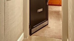Un couloir avec une porte fermée, un tableau encadré accroché au mur, et on aperçoit une pièce avec un lit et une fenêtre.