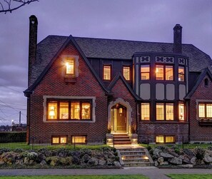 A large, two-story house with a prominent chimney, a well-lit entrance, and a stone foundation. - Suite I: Private Studio in our classic home near Woodland Park Zoo (Seattle)