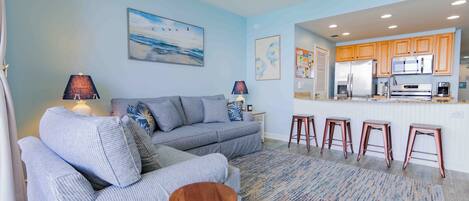A living room with a sofa, a coffee table, and a kitchen area with a refrigerator and bar stools.