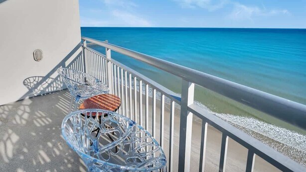 Een balkon met uitzicht op een helderblauwe oceaan, witte leuningen en twee glazen stoelen.