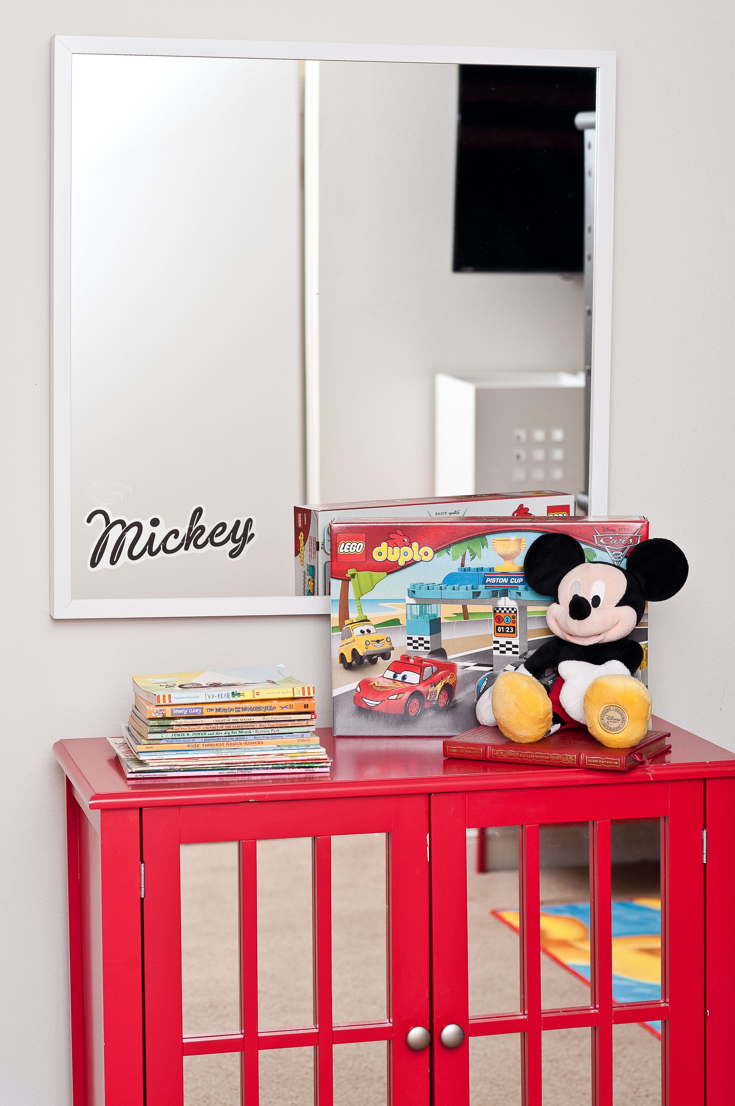 Ein roter Schrank mit Spiegel darüber, ein Stapel Bücher und ein Mickey Mouse Stofftier darauf.