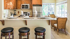 Une cuisine avec des meubles de rangement en bois, un réfrigérateur en acier inoxydable, un four à micro-ondes et une grande fenêtre. On y trouve trois tabourets de bar et un fauteuil en osier.