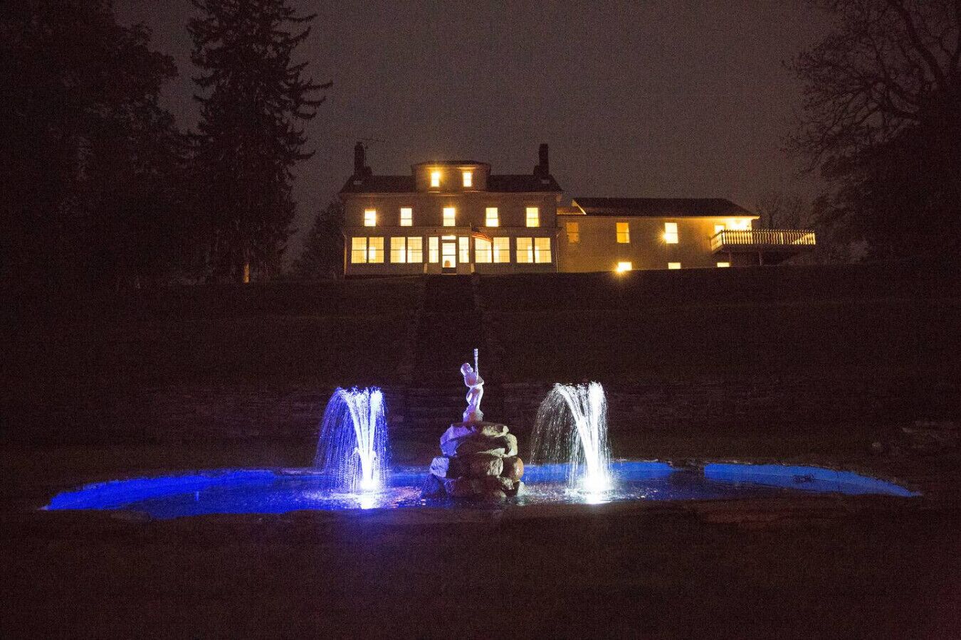 Vista nocturna de un edificio con varias ventanas iluminadas y una fuente con chorros de agua.