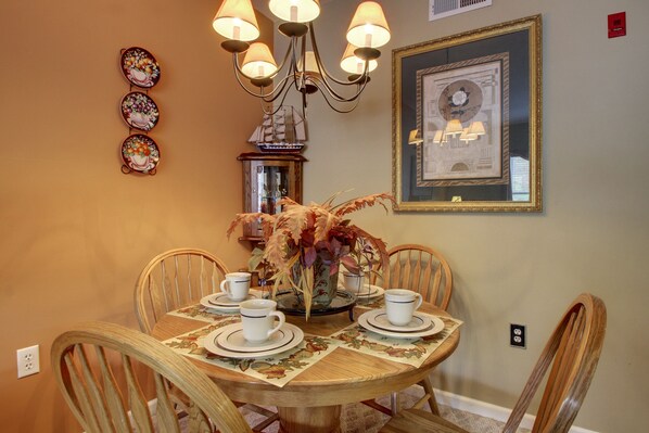 A dining area with a round wooden table, white plates, and matching cups. A chandelier hangs above, and a framed picture is on the wall.