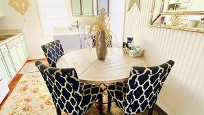 A dining area with a round table, four patterned chairs, a vase with dried plants, a ceiling fan, and a kitchen in the background. - 2 Minute walk to the secluded beaches of Fort Morgan! Pet friendly! Quiet Space! (Gulf Shores)