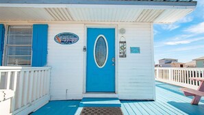 A small, single-story building with a blue door and a white exterior. There is a sign above the door with text, a small window with a decorative glass design, and a wooden deck with a bench. - 2 Minute walk to the secluded beaches of Fort Morgan! Pet friendly! Quiet Space! (Gulf Shores)