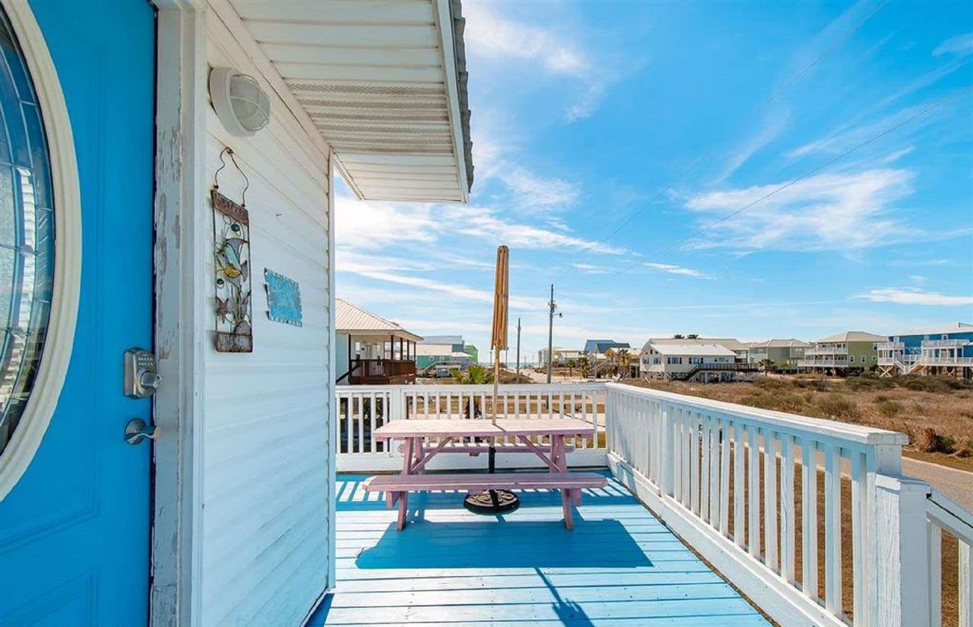 A blue wooden deck with a picnic table, a white railing, and a clear view of the beach and houses in the background.