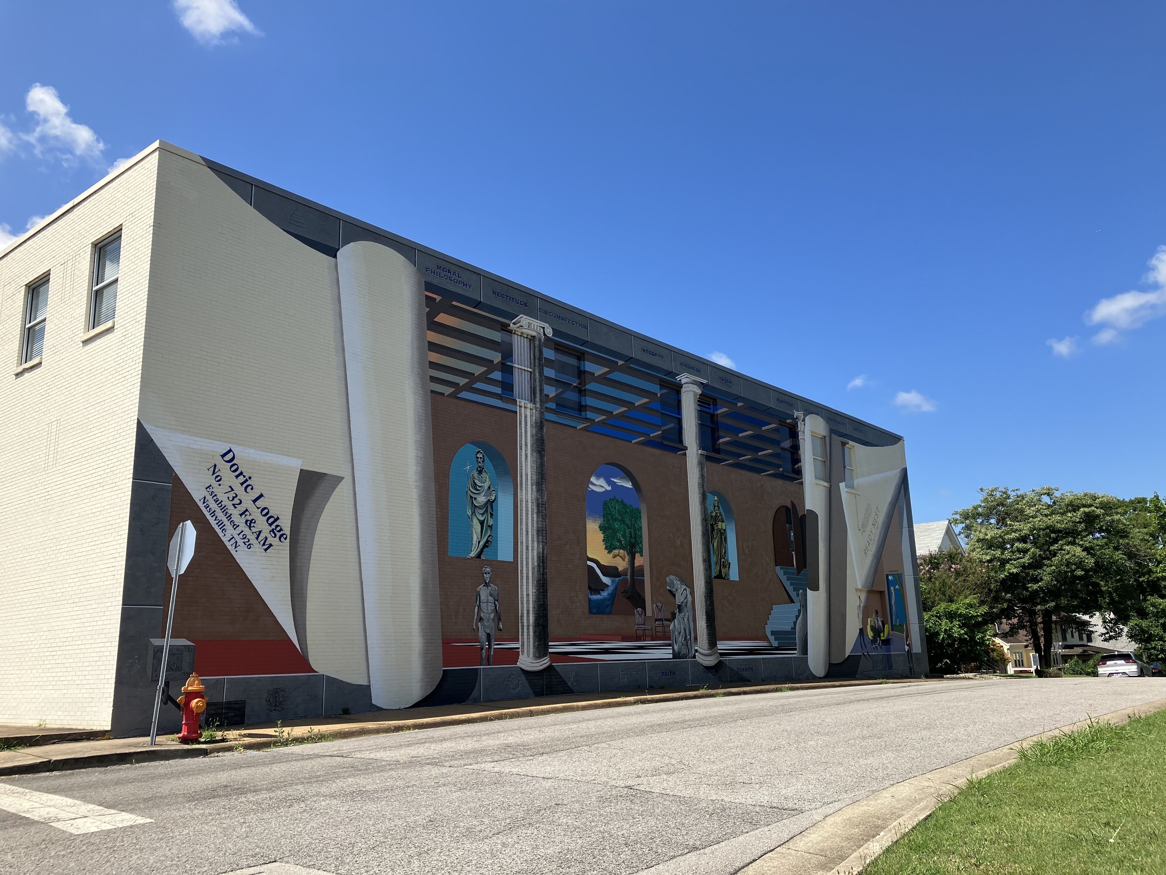 A building with a mural of classical figures and columns, a fire hydrant, and a clear blue sky.