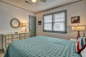 Een slaapkamer met een bed, een dressoir, een raam met jaloezieën, een spiegel en een wandlamp.