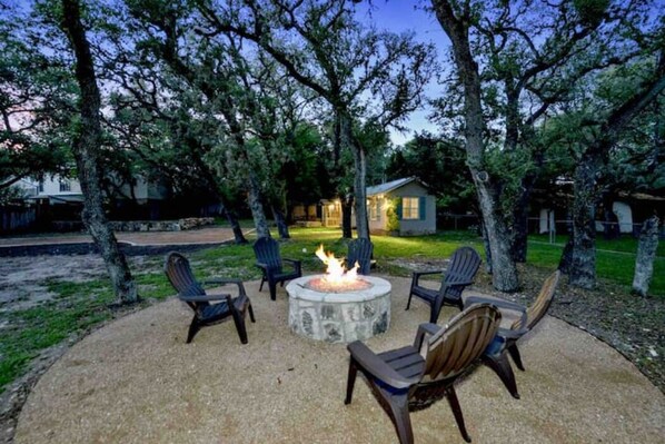 A fire pit surrounded by chairs in a backyard with trees and a house in the background.