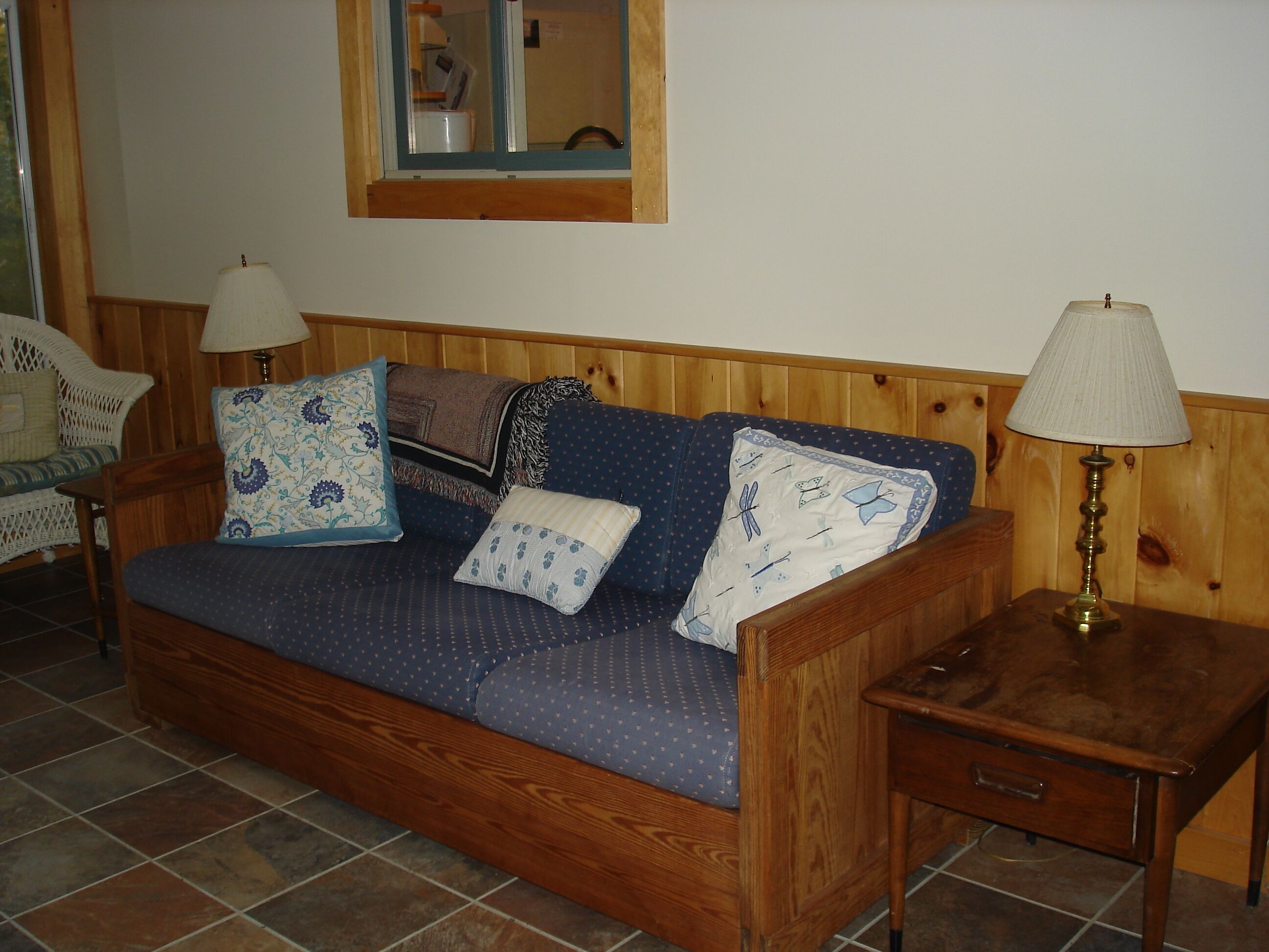 A wooden sofa with patterned cushions, a small wooden table with a lamp, and a wicker chair in a room with wooden paneling and tiled flooring.