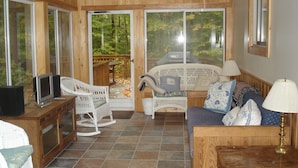Una sala de estar acogedora con televisor, muebles de mimbre y vistas a una terraza de madera y árboles.