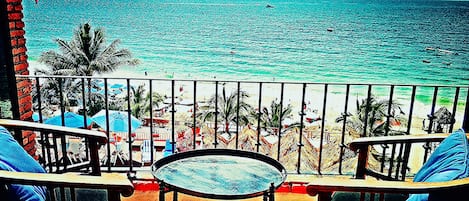 A balcony with a table and chairs, overlooking a beach with palm trees and a clear view of the ocean.