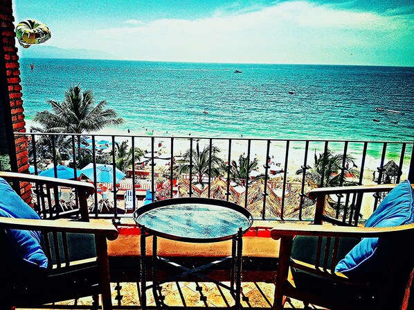A balcony with a table and chairs, overlooking a beach with palm trees and a clear view of the ocean.