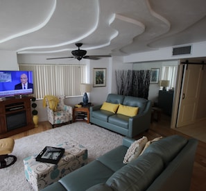 Un salon avec un canapé bleu, un pouf à motifs, un téléviseur et un ventilateur de plafond.