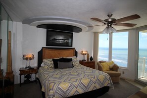Une chambre dotée d’un grand lit, d’un ventilateur de plafond, d’une table de chevet avec une lampe, d’un fauteuil avec un coussin et offrant une vue sur l’océan grâce à de grandes fenêtres.