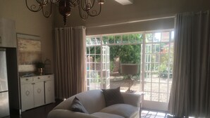 A living room with a sofa, a chandelier, a cabinet, and a large window with curtains. - Solar powered. Large Garden Apartment, Central & Secure Neighbourhood. (Johannesburg)