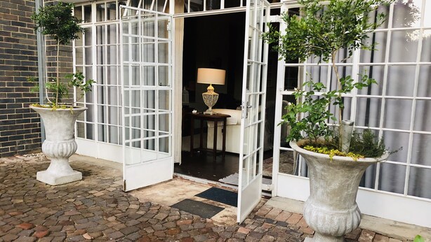 A building entrance with two large potted plants, a white door, and a brick wall. - Solar powered. Large Garden Apartment, Central & Secure Neighbourhood. (Johannesburg)