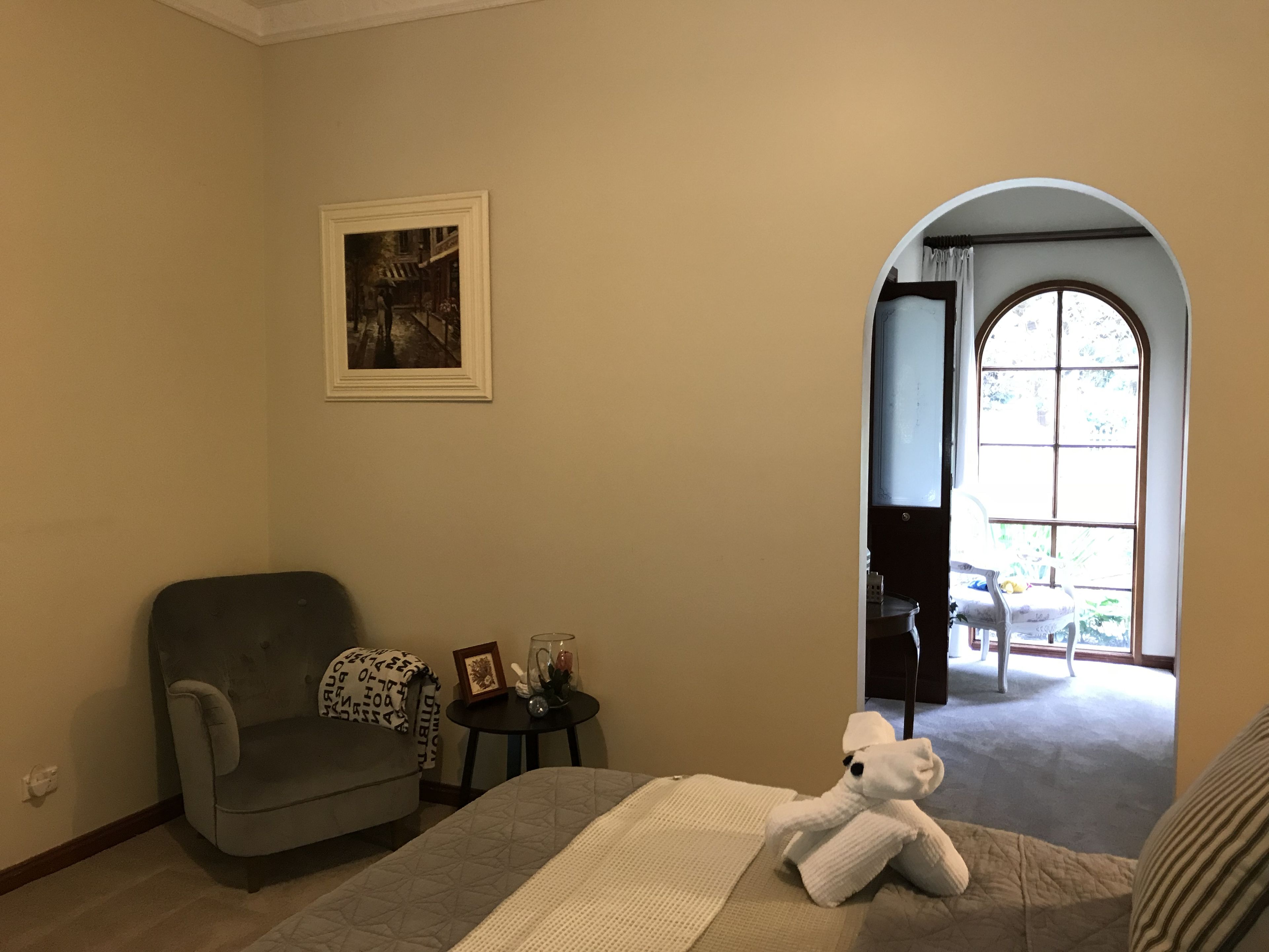 Habitació d'hotel amb una butaca grisa, una taula petita amb un quadre i un got, un llit amb llençols blancs, i una finestra gran d'arc amb vista a l'exterior.