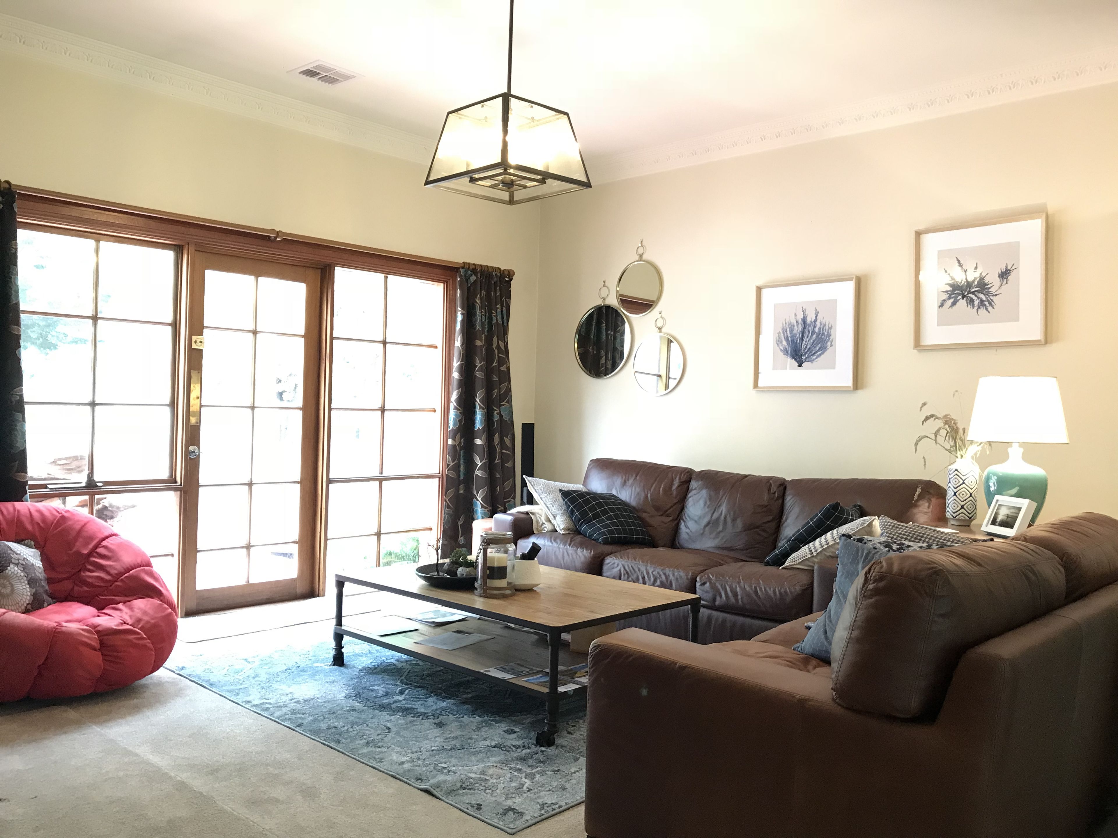 A living room with a brown leather sofa, a red bean bag chair, a coffee table, and a large window with curtains.